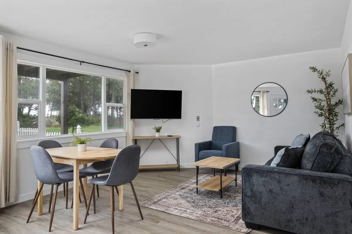 A cozy living and dining area with a dining table, comfortable sofa, television, large window, and lush indoor plants. The arrangement looks ideal for family meals, relaxed evenings, and watching sunsets together.
