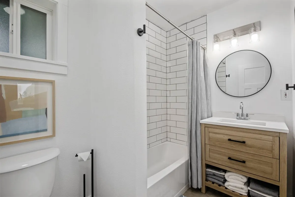 A bright, modern bathroom featuring a wooden vanity, circular mirror, a spacious shower area, and framed artwork. The natural light and fresh fixtures emphasize the clean, contemporary updates in the space.
