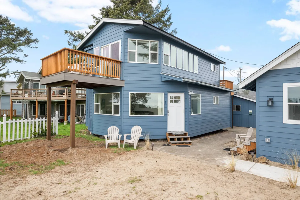 Two white chairs on a wooden balcony facing the blue siding of the house, with an inviting yard in front. The setting is relaxed, suggesting moments of morning coffee, sunset views, or evening gatherings around a fire pit, with strong indoor-outdoor flow.