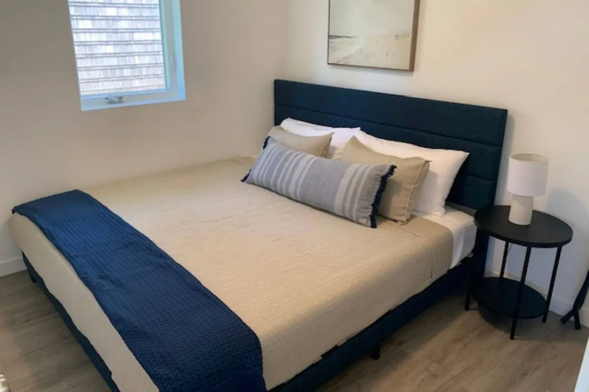 A tranquil bedroom space with a comfortable king bed, accent pillows, a nightstand, and relaxing artwork. The gentle lighting and soft tones create an inviting atmosphere ideal for relaxation and sleep.