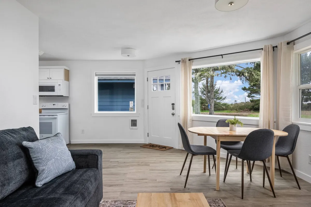 A welcoming open-concept living area with a comfortable sofa, inviting dining table, and large windows that flood the space with natural light. The space is styled with modern furnishings and subtle coastal decor, creating a bright and airy atmosphere. The windows offer glimpses of the beach just outside.