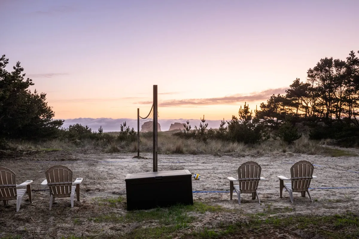 A tranquil exterior scene featuring Adirondack chairs arranged around a communal fire-pit, a sandy volleyball court with a volleyball net, and the backdrop of a glowing sunset. The setting encourages both relaxation and active play, with the communal spaces positioned to take advantage of the surrounding natural views.