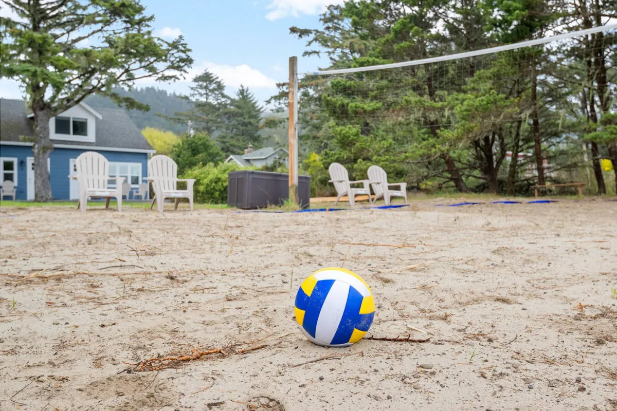 A fun and inviting view of the sand volleyball court, with a volleyball net, Adirondack chairs for spectators, and surrounding trees. The sandy area and clear open sky create an energetic, beachy feel ideal for group activities and relaxation.