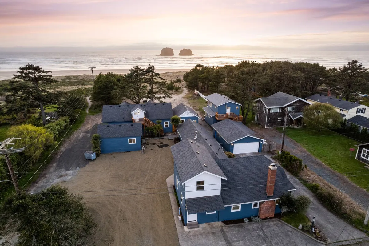 This image features a scenic beachfront with houses overlooking the sand, the iconic Twin Rocks formation visible in the background, and a glowing sunset over the ocean. Children and families are seen playing and gathering on the shore, with sandcastles and the peaceful ambiance of the beach setting.