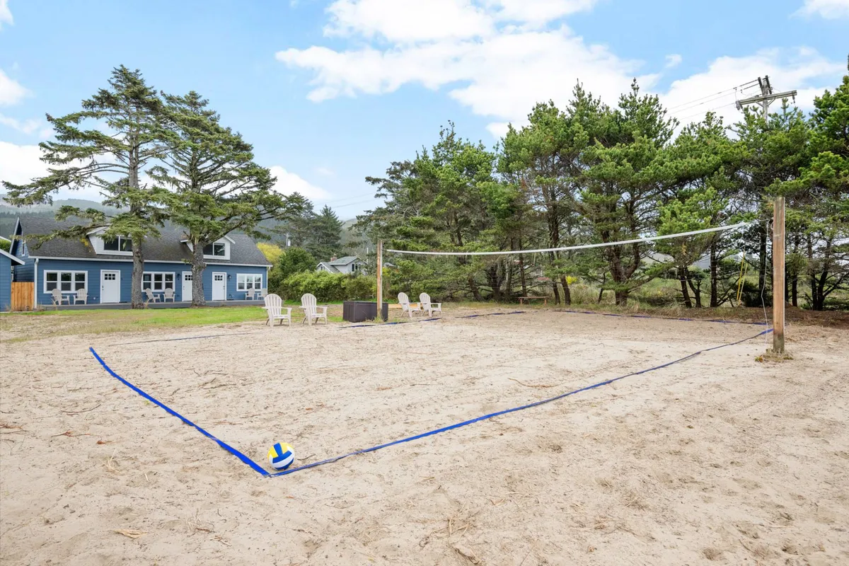 The image shows a lively beach scene centered around a sand volleyball court with a net, surrounded by outdoor seating, trees, and a wide blue sky. Families and friends are seen playing and laughing together, while others relax nearby, with the sense of community and seaside recreation.
