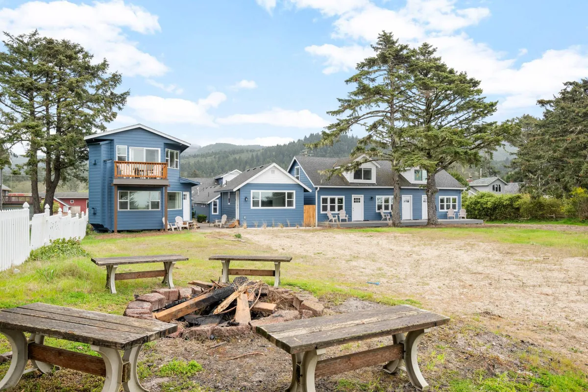A cozy outdoor gathering area with a central fire pit, surrounded by blue cottages and a large tree. The setting is perfect for group gatherings, storytelling, or stargazing, with a friendly and communal mood.