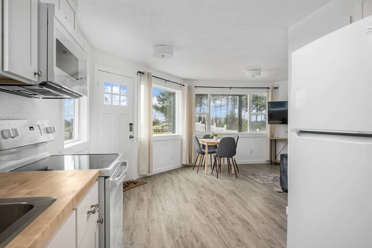 A bright kitchen and dining area featuring a kitchen island, large windows, and a dining table. The space feels cozy and functional, with ample natural light and a layout perfect for socializing, cooking, and enjoying family game nights.