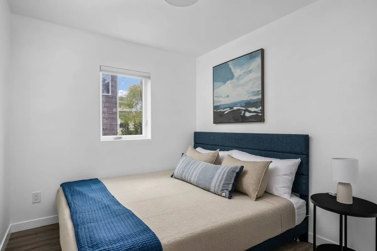 A serene bedroom with a neatly made bed, calming artwork above the headboard, and a window allowing in soft natural light. The room features soothing decor and gentle lighting that creates a tranquil ambiance, perfect for restful nights.