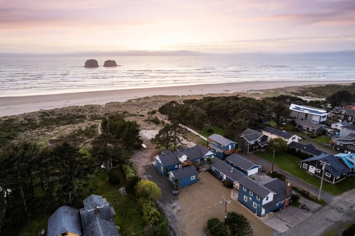 The image showcases a serene view of Rockaway Beach with soft sand stretching towards the iconic Twin Rocks Arch formations in the Pacific Ocean. Charming coastal houses are nestled along the shore, and the tranquil scene is bathed in warm sunset hues, inviting guests to imagine strolling the shoreline or gathering around a beach fire as the day ends.